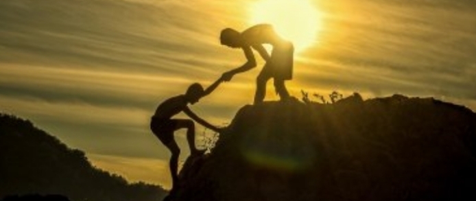 Silhouette of one person helping another climb a rocky hill at sunset.